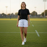 A woman wearing Ruggers Rugby Supply Auckland white rugby shorts with stretch panels on a green sports field.